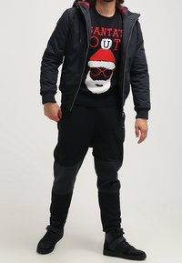 Schwarze gepolsterte Jacke mit roten Akzenten, schwarzer Weihnachts-Pullover mit Santa-Grafik, schwarze Jogginghosen mit strukturierten Paneelen und schwarze Sneakers.