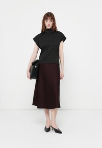Top nero a collo alto con maniche a campana abbinato a una gonna midi in satin bordeaux. Il modello tiene una minaudière nera e indossa mules con tacco neri.