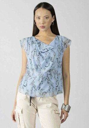 Donna che indossa una blusa floreale azzurro chiaro con collo arricciato, pantaloni cargo beige, braccialetto d'argento e orecchini a cerchio su uno sfondo semplice.