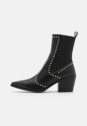 Bottine en cuir noir avec bout pointu, talon épais, panneau élastique latéral et clous dorés décoratifs soulignant les coutures.
