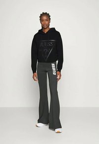 Kobieta ubrana w czarną bluzę z kapturem marki Guess i ciemnoszare spodnie dzwony z białym logo Guess, stojąca na tle jednolitego jasnego tła.