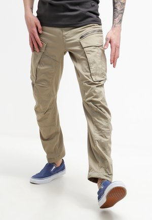 Person wearing beige cargo pants with multiple pockets and blue slip-on sneakers, standing with one foot forward on a white background.