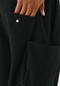 Pantaloni neri realizzati in tessuto testurizzato, dotati di tasche laterali angolate con dettaglio a scatto, che presentano un design funzionale e pratico.