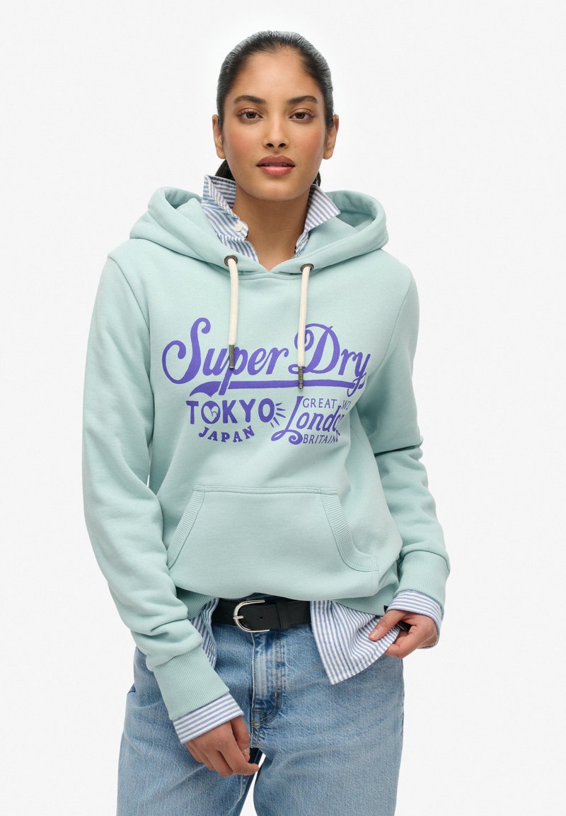 Jasno miętowy hoodie z przednią kieszenią, kremowymi sznurkami i fioletowym napisem; nałożony na niebieską w białe paski koszulę z kołnierzykiem.
