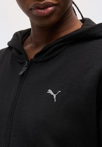 Κοντινό πλάνο ενός ατόμου που φοράει μαύρο φούτερ με φερμουάρ και μικρό λευκό λογότυπο Puma στο αριστερό μέρος του στήθους.