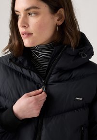 Casaco puffer preto com fecho à frente e capuz ajustável. Apresenta um pequeno logótipo da Levi's no peito e punhos canelados. Camisola de gola alta às riscas por baixo.