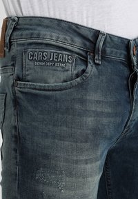 Jeans de mezclilla en un color azul desgastado con una superficie texturizada. Presenta un parche con el logotipo, diseño de cinco bolsillos y detalles desgastados.
