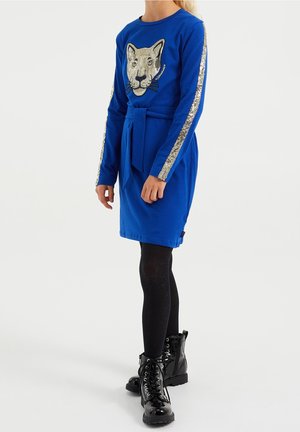 Enfant portant une robe bleue à manches longues avec une ceinture nouée, visage de lion en sequins sur le devant, bandes argentées en sequins sur les manches, collants noirs et bottes noires.