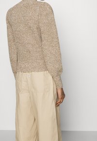 Strickpullover in hellbraun mit strukturiertem Gewebe und langen, gepufften Ärmeln; kombiniert mit lockeren beigen Hosen mit Gesäßtaschen.