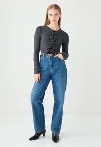 Grijze lange mouwtop met een gerimpelde knopenlijst; hoog-waist blauwe jeans; zwarte puntige hakken. Eenvoudige achtergrond.