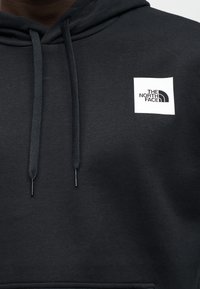 Μαύρο φούτερ με κουκούλα και υφή, με κορδόνι στη λαιμόκοψη και άσπρο τετράγωνο μπαλώμα με το λογότυπο "THE NORTH FACE" στο στήθος.
