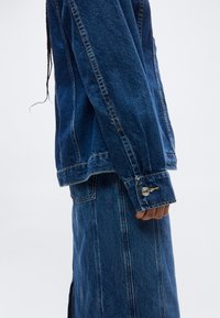 Άτομο που φοράει μπλε τζιν σακάκι και τζιν φούστα στο ίδιο χρώμα, στέκεται πλευρικά με το αριστερό του χέρι χαλαρό δίπλα στο σώμα.