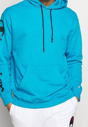 Person trägt einen hellblauen Hoodie mit schwarzen Kordeln und weiße Hose mit schwarzem und rotem Logo, Hände in den Taschen.
