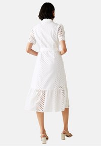 Kobieta w białej sukience midi z koronką, z krótkimi rękawami, paskiem w talii i falbaniastym dołem, w beżowych sandałach na szerokim obcasie, stojąca tyłem.