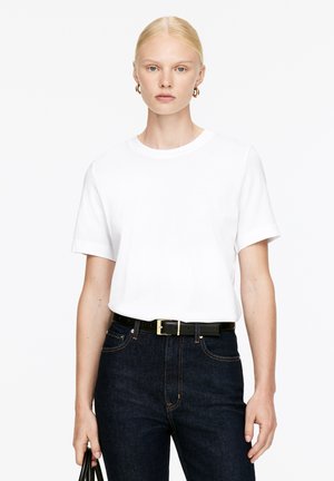 T-shirt en coton blanc avec un col rond et des manches courtes retroussées, associée à un jean en denim foncé et une ceinture noire avec une boucle dorée.