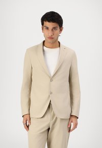 Beige sako na míru s dvěma knoflíky, klopami, hladkou texturou a přiléhavým střihem. Nosit na bílé tričko.