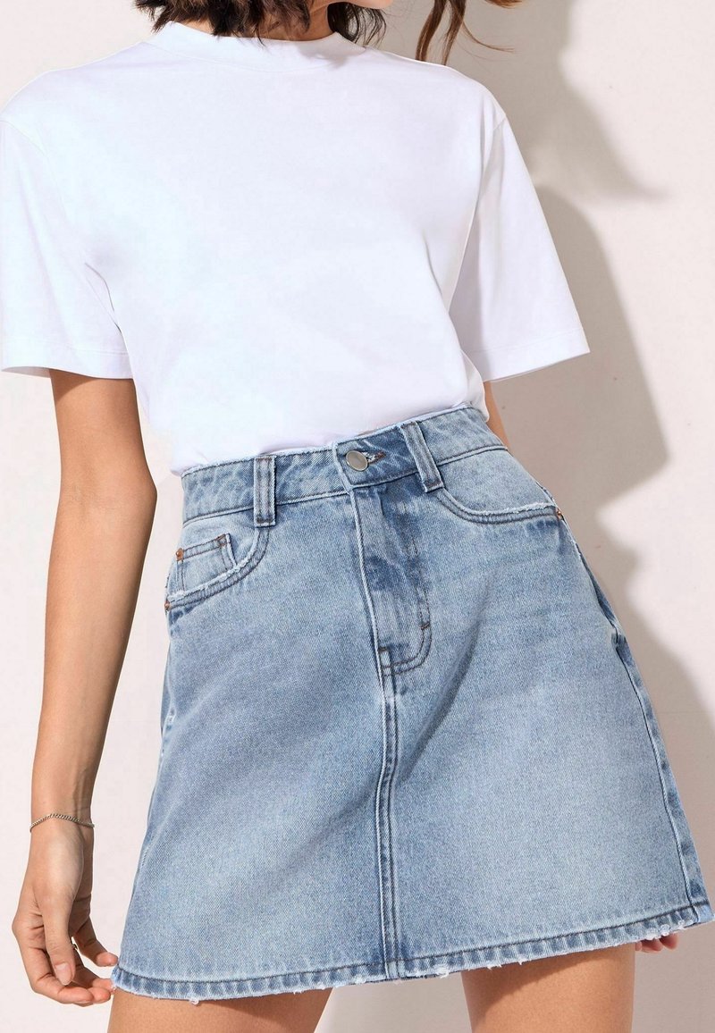 Femme portant un t-shirt blanc uni à manches courtes, rentré dans une mini-jupe en denim bleu clair taille haute avec poches avant et fermeture par boutons.