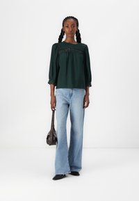 Blusa verde con texture, maniche a sbuffo e dettagli in pizzo; abbinata a jeans azzurri a zampa d'elefante e una piccola borsa a tracolla marrone.