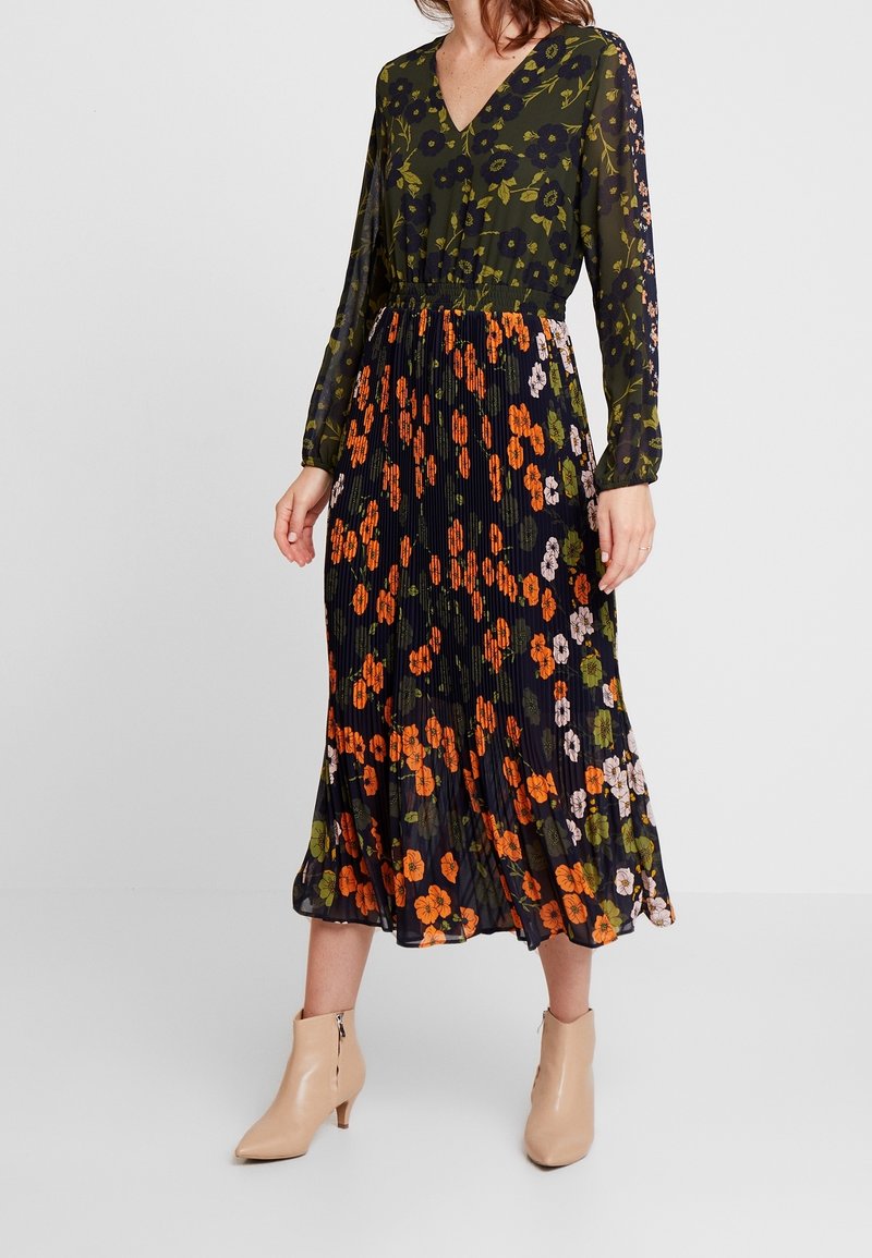 Femme portant une robe fleurie à manches longues avec un haut vert et une jupe noire plissée avec des fleurs orange, associée à des bottines beiges.