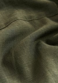 Tissu en lin vert foncé avec une texture lisse, présentant des motifs de tissage subtils et une couture surpiquée, créant de doux plis.