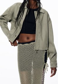 Olijfgroene spijkerjasje met drukknopen en een losse pasvorm, gecombineerd met een zwart cropped top en een gestructureerde, zwarte en olijfgroene netrok.