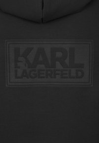 Svart hoodie med en upphöjd, tonad logotryck av "KARL LAGERFELD" tillsammans med en siluett av en figur, tillverkad av mjukt bomullsmaterial.