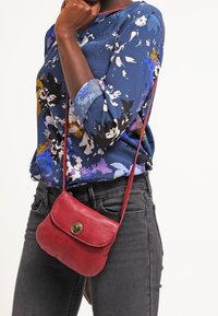 Röd läder crossbody-väska med rundad form, framfällbar stängning och en framträdande gul knapp. Bärs tillsammans med en blommig blå blus och svarta jeans.