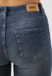 Jeans de mezclilla azul oscuro con una textura suave, que presentan una etiqueta de cuero en la cinturilla y dos bolsillos traseros con acentos de costura.