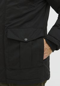 Črna, izolirana jakna z žepom na klapni in stranskim žepom za roke. Ima gladko teksturo in prilegajoč se manšet. Gumbi so nerezljivi.