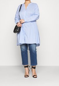 Blauw-wit gestreepte jurk met kraag en knopen, gecombineerd met blauwe jeans en zwarte haksandalen, en een zwarte schoudertas.