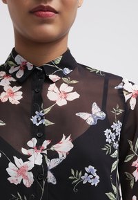 Blouse noire à imprimé floral en matière transparente, présentant des fleurs blanches et bleues ainsi que des papillons bleu clair. Col chemise avec boutons sur le devant.