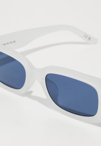 Lunettes de soleil blanches avec une finition mate et des verres teintés en bleu. Monture rectangulaire élégante avec des branches courbées et un design minimaliste.