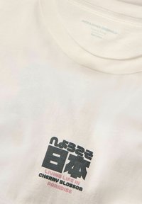 Bela majica s okroglim ovratnikom z črnimi in roza napisi, vključno z napisom "ŽIVETI ŽIVLJENJE V RAJU CVETOČIH BRESKEV" in azijskimi znaki.