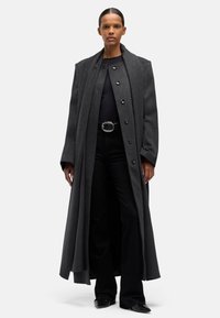 Długi, ciemnoszary wełniany płaszcz z wysokim kołnierzem, o oversize'owym kroju, z sześcioma czarnymi guzikami z przodu, zestawiony z czarnymi spodniami i butami.
