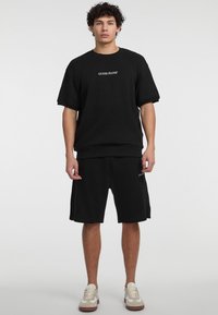 Sæt bestående af en sort bomulds sweatshirt og shorts med et lille hvidt "GUESS JEANS" logo, afslappet pasform og parret med beige og hvide sneakers.