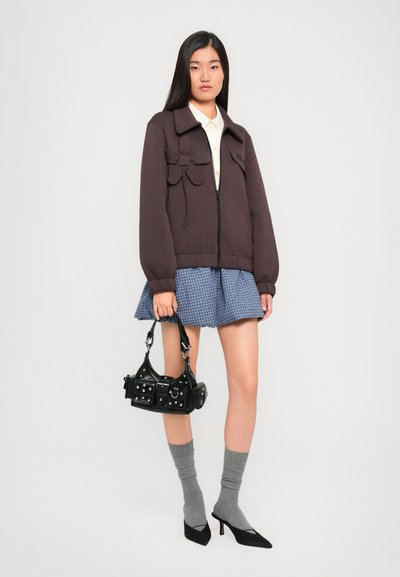 Chaqueta de cremallera marrón oscuro con adornos florales, camisa clara, falda a cuadros azul, calcetines grises y bolso negro con detalles de metal.