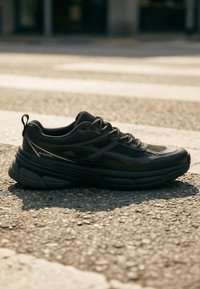 Baskets de sport noires avec une semelle épaisse et un design à lacets, placées sur un asphalt texturé près d'un passage piéton sous la lumière du soleil.