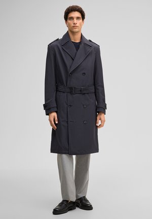 Man met een donkerblauwe double-breasted trenchcoat met riem, lichtgrijze broek en zwarte loafers, staand tegen een effen grijze achtergrond.