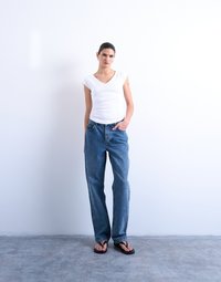 Γυναίκα με σκούρα μαλλιά, φορώντας λευκό κοντομάνικο τοπ, μπλε τζιν και μαύρα σανδάλια, στέκεται μπροστά σε έναν απλό λευκό τοίχο με το ένα χέρι στην τσέπη.