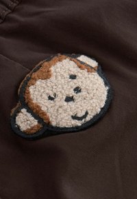 Brauner Stoff mit einem strukturierten, runden Äffchen-Patch in Beige und Braun, umrissen in Schwarz, mit einem lächelnden Gesicht und zwei Ohren.