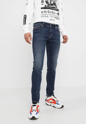 Uomo che indossa jeans slim-fit blu scuro, maglietta bianca a maniche lunghe con grafica e sneakers bianche con accenti rossi e blu, in piedi su pavimento bianco.