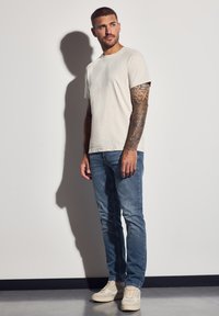 Jasnoszara koszulka z okrągłym dekoltem i krótkimi rękawami, w połączeniu ze średnio niebieskimi dżinsami i beżowymi trampkami, na ramionach widoczne tatuaże.