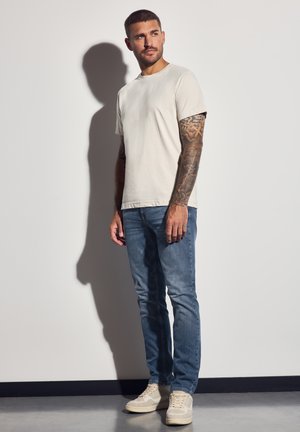 Camisa de tirantes gris claro con cuello redondo y mangas cortas, combinada con jeans azules medios y zapatillas beige, mostrando tatuajes visibles en los brazos.