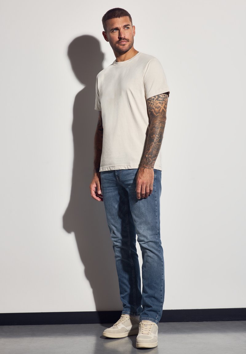 Jasnoszara koszulka z okrągłym dekoltem i krótkimi rękawami, w połączeniu ze średnio niebieskimi dżinsami i beżowymi trampkami, na ramionach widoczne tatuaże.