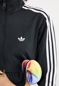 Veste noire zippée avec détail à trois bandes blanches, présentant un logo brodé. Un tissu coloré apparaît par les manches, mettant en avant un motif dégradé.