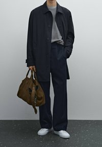 Marineblauen Mantel über grauem Hemd, weite marineblaue Hose, weiße Sneakers und eine braune Lederhandtasche mit dunklen Akzenten und einem Reißverschlussdetail.