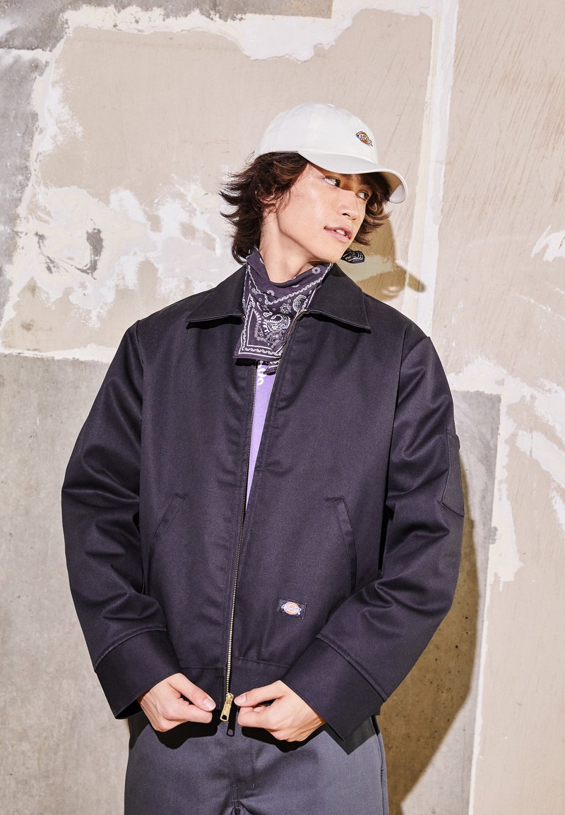 Chaqueta negra con cremallera y cuello, cuenta con bolsillos laterales y un pequeño parche con logo. Lleva puesta sobre una camisa morada, combinada con un pañuelo estampado.