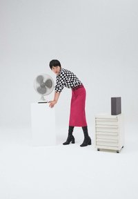 Blusa a pois bianca e nera, gonna a tubino fucsia, stivali neri fino al ginocchio. In piedi accanto a un ventilatore a rete su un piedistallo, con un'unità di stoccaggio nelle vicinanze.