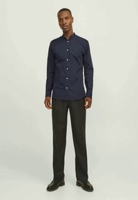 Tume sinine nööpidega särk, seisva kraega, kohandatud sobivuse ja pikkade varrukatega, kombineeritud tumedate pükste ja mustade kingadega. Sile kangateks.