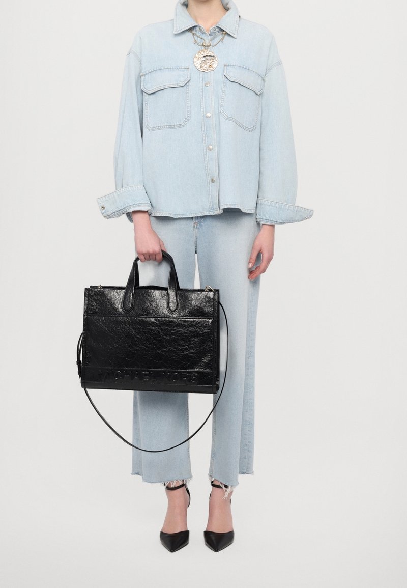 Personne portant une veste en jean bleu clair et un jean, tenant un grand sac à main en cuir noir et portant des talons hauts noirs pointus.